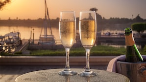 Dos copas de champán sobre una mesa con una botella, frente a una vista del lago y barcos al atardecer.