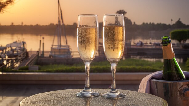 Two champagne flutes on a table with a bottle of champagne, set against a sunset view of a lake and boats. - Jolie Ville Hotel & Spa Kings Island Luxor (Luxor)