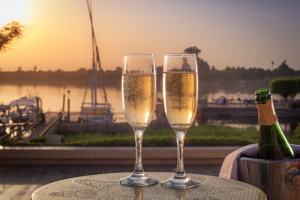 Dos copas de champán sobre una mesa con una botella, frente a una vista del lago y barcos al atardecer.