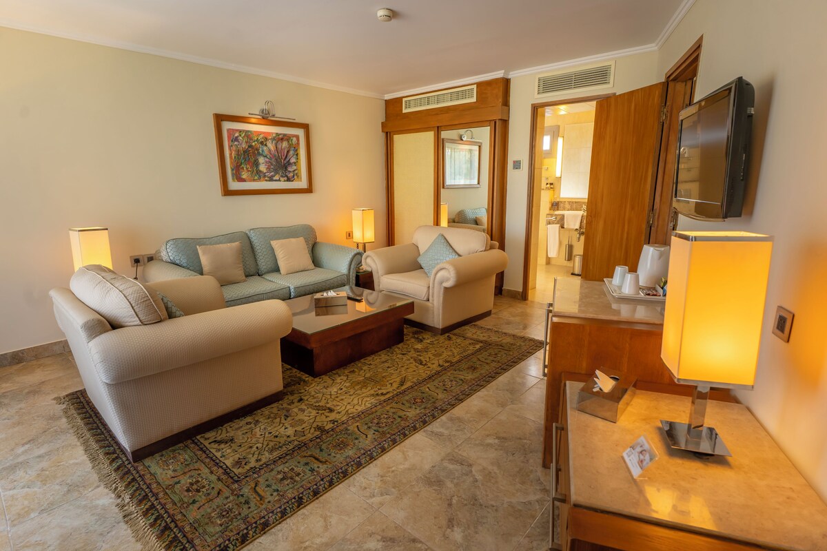 A living room with a sofa set, a coffee table, a TV, and a kitchen area visible through an open door.