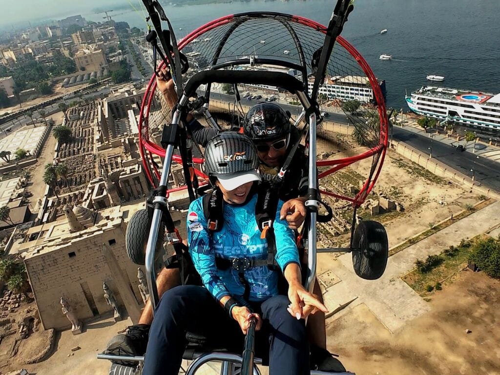 A person in a blue jacket and helmet, seated in a red and black chair with a view of ancient ruins and a cruise ship in the background.