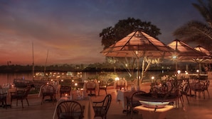 Un área de comedor al aire libre con mesas y sillas, sombrillas y vistas al agua al atardecer.