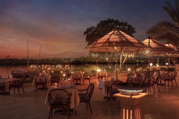 Un área de comedor al aire libre con mesas y sillas, sombrillas y vistas al agua al atardecer.