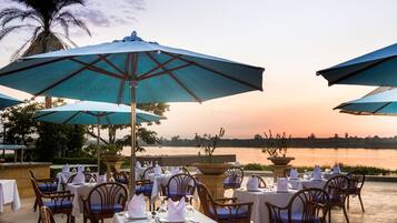 An outdoor dining area with tables and chairs, large umbrellas, and a view of a body of water at sunset.