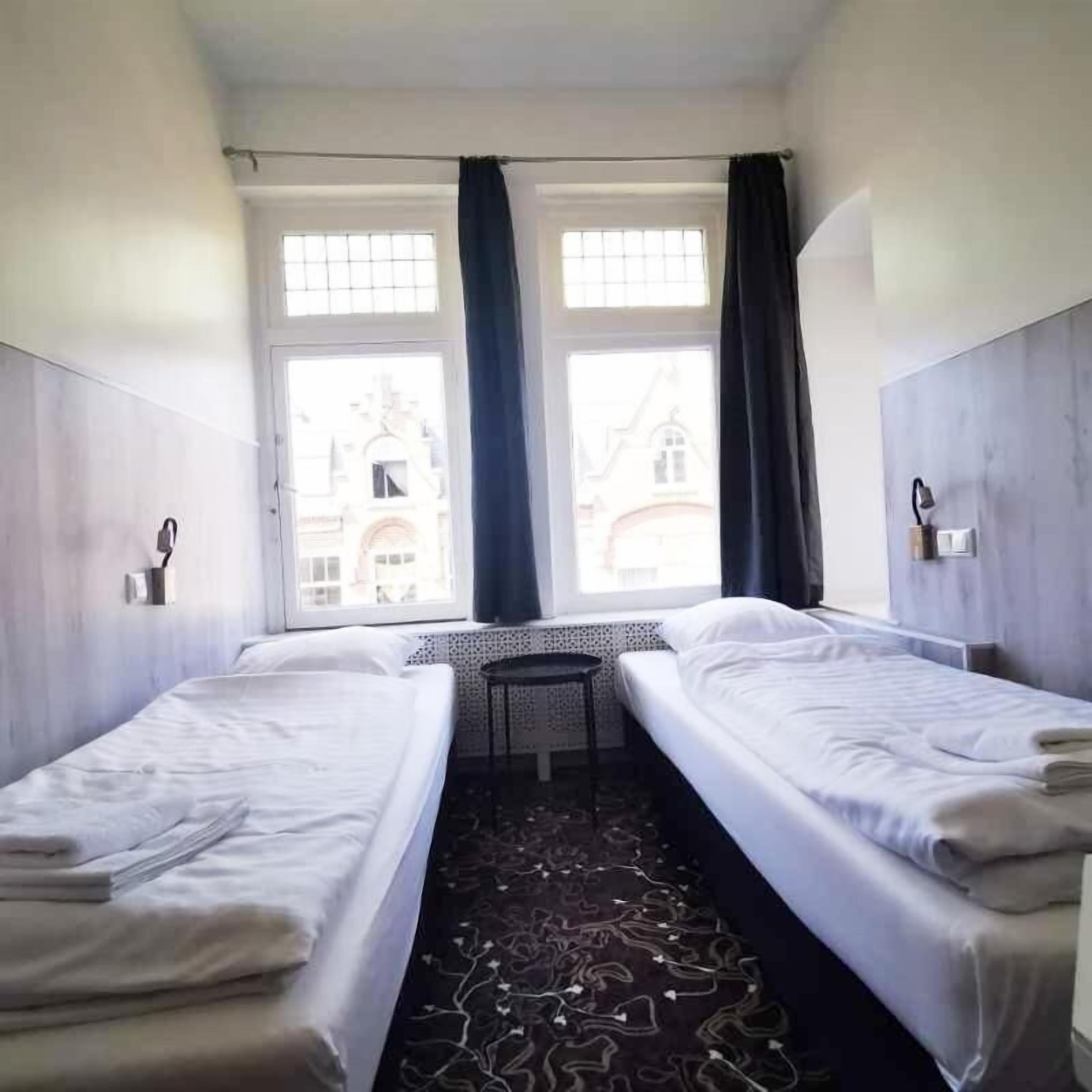 A hotel room with two single beds, a small round table, and a view of a building with a clock tower through large windows.