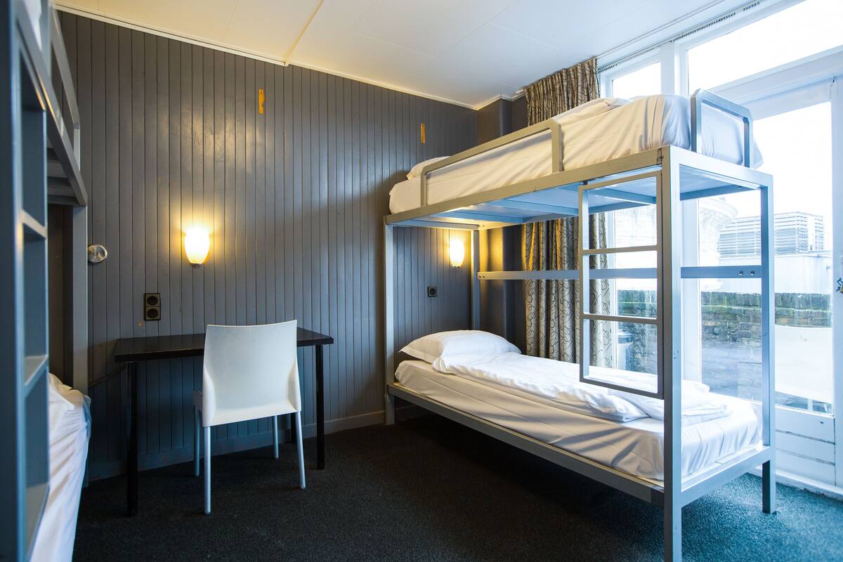 A bunk bed room with a desk and chair, a window with curtains, and a wall-mounted light.