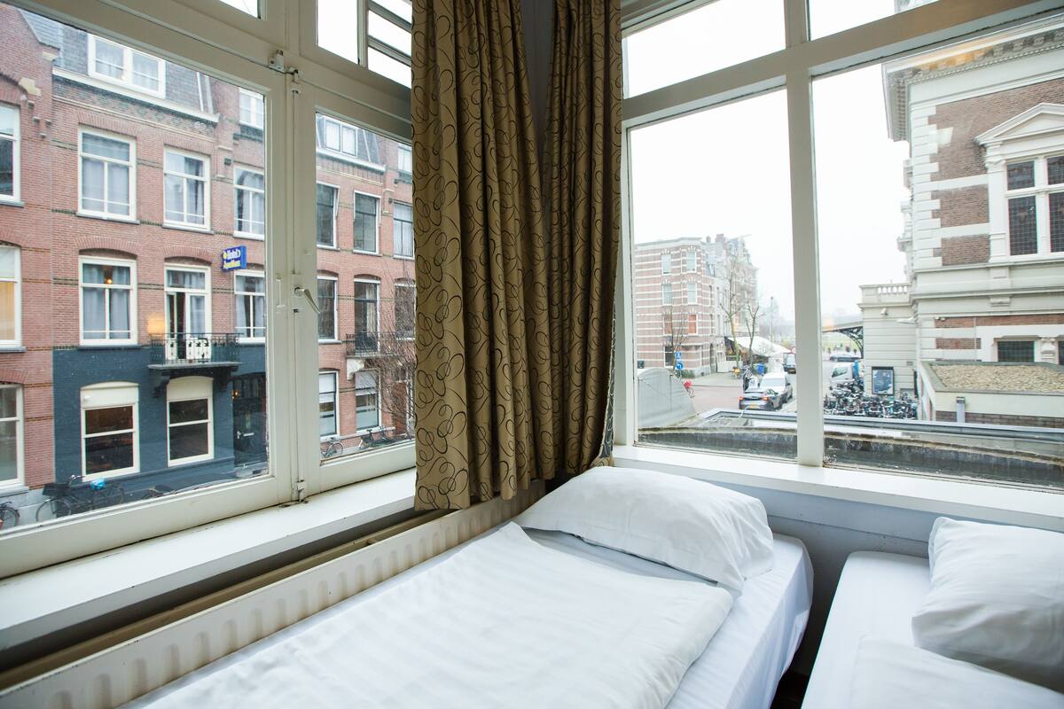 A hotel room with two beds, a large window with curtains, and a view of the cityscape.