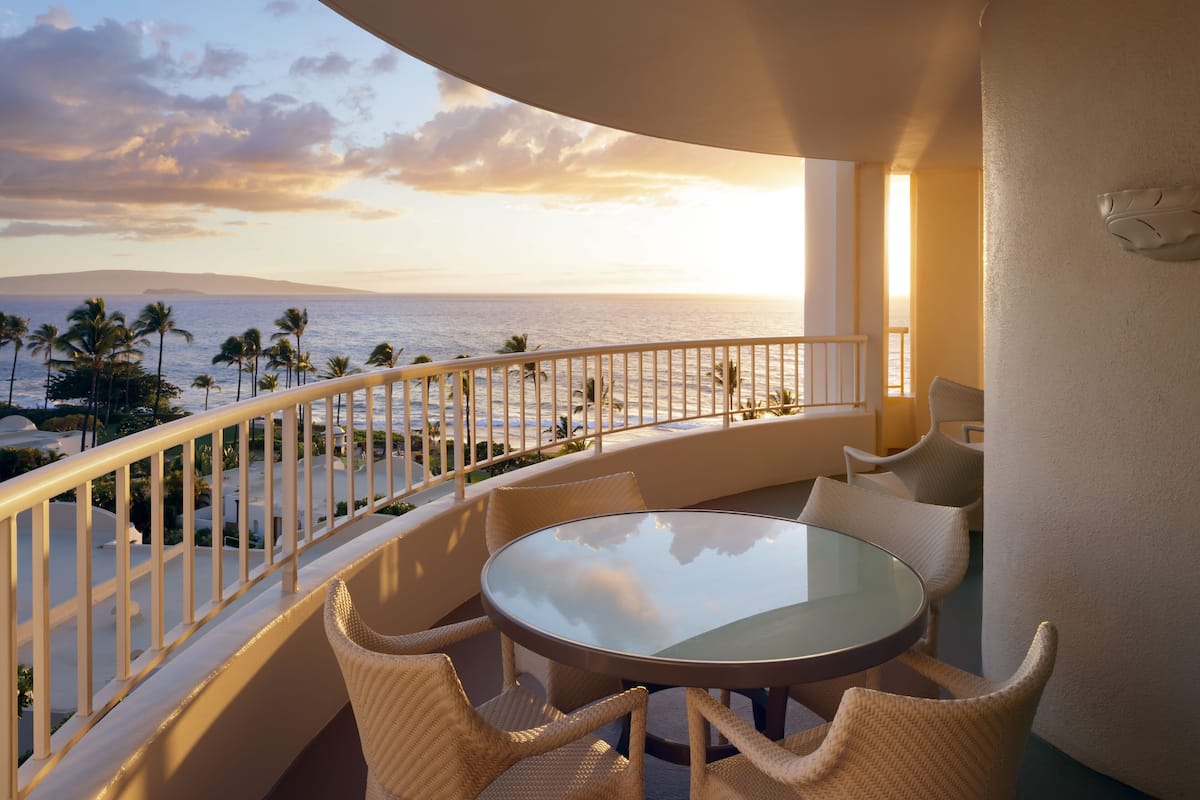 A balcony with a glass table and wicker chairs, overlooking a beach with palm trees and the ocean.
