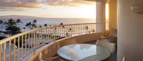 Un balcon doté d’une table en verre et de chaises en osier, offrant une vue sur une plage bordée de palmiers et sur l’océan.