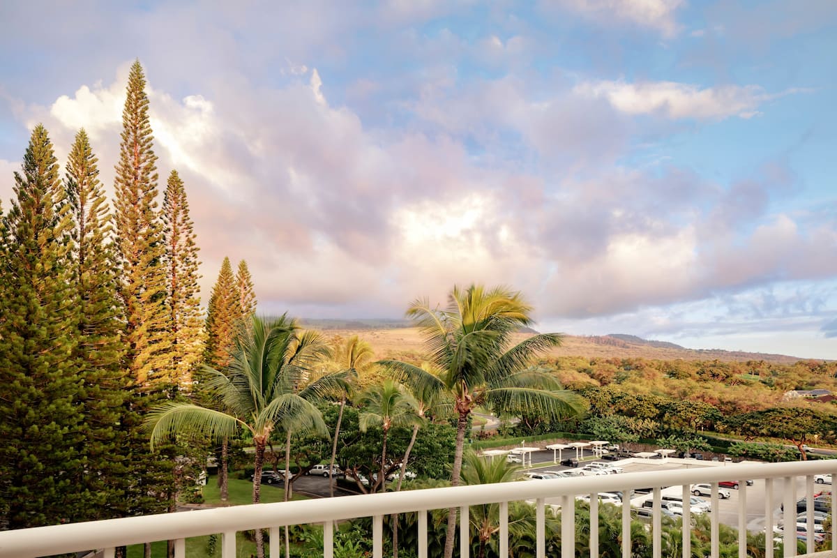 A view from a balcony overlooking a parking area with palm trees and coniferous trees, with a mountainous landscape in the background.