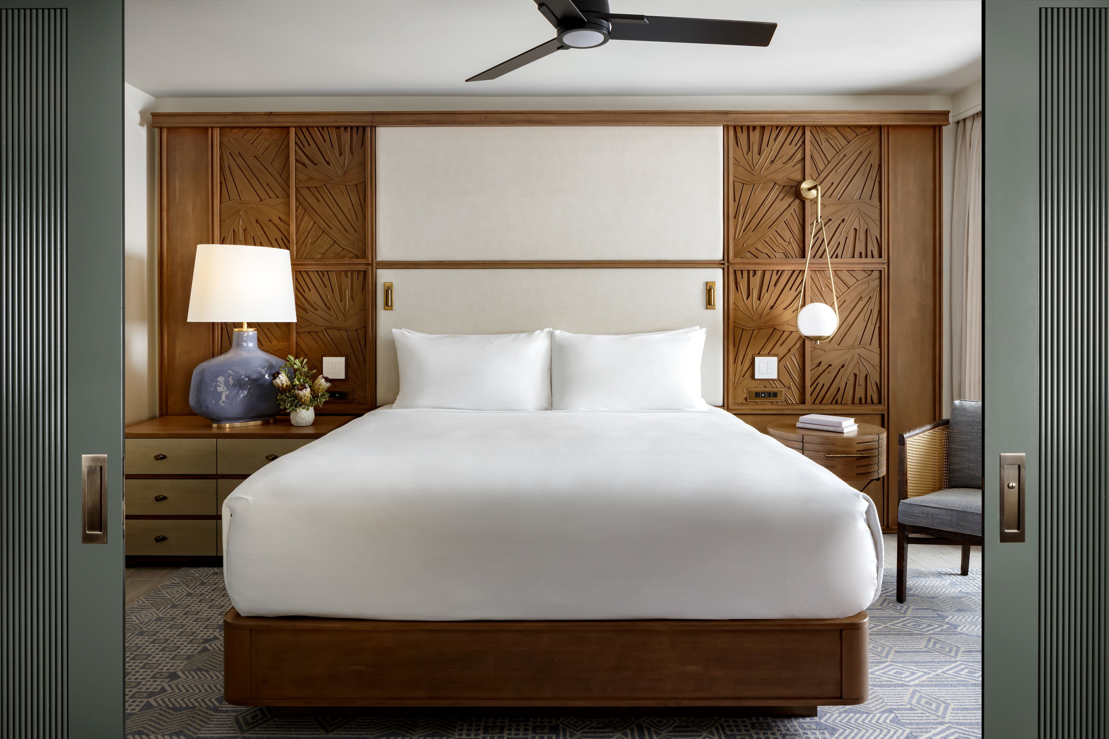 A large bed with white linens, a ceiling fan, wooden paneling on the walls, a nightstand with a lamp, and a chair.