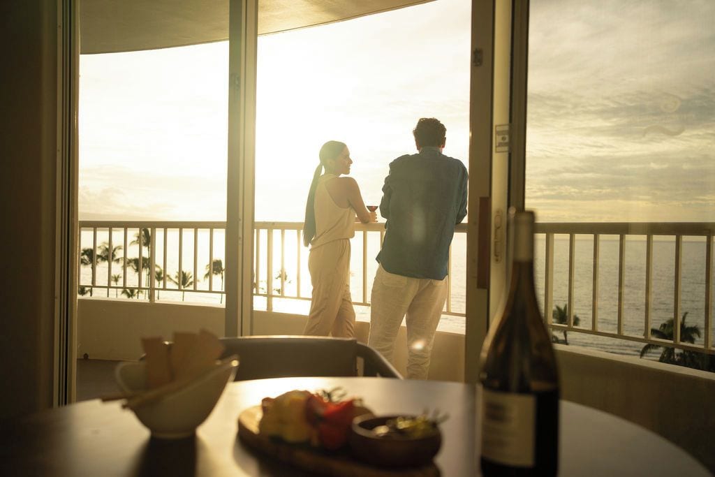 A couple standing by a balcony with a view of the sea, a table with food and a bottle of wine, and a railing.