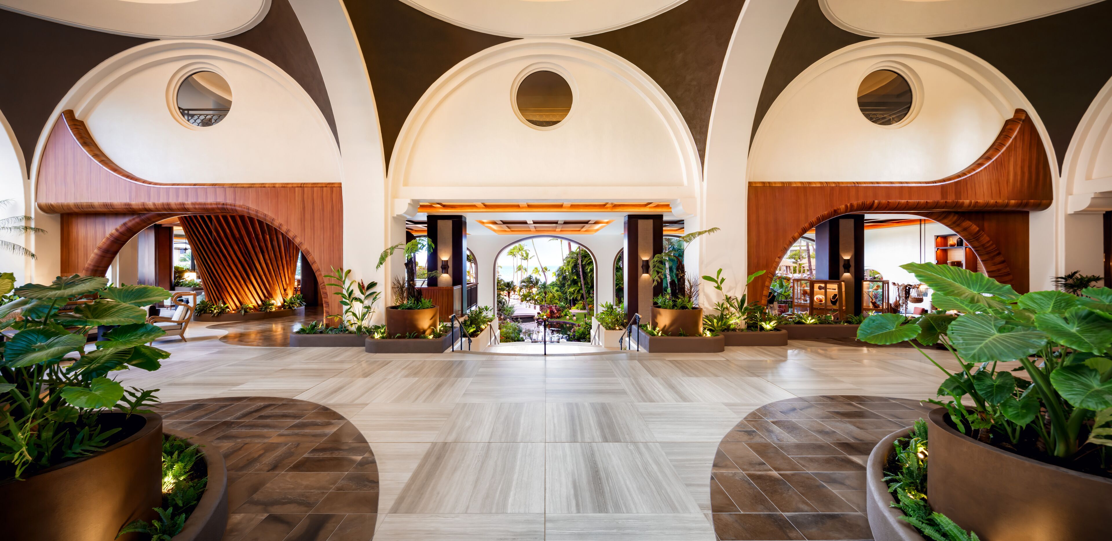 A spacious lobby with arched ceilings, large planters with tropical plants, and a central open area leading to a seating area.