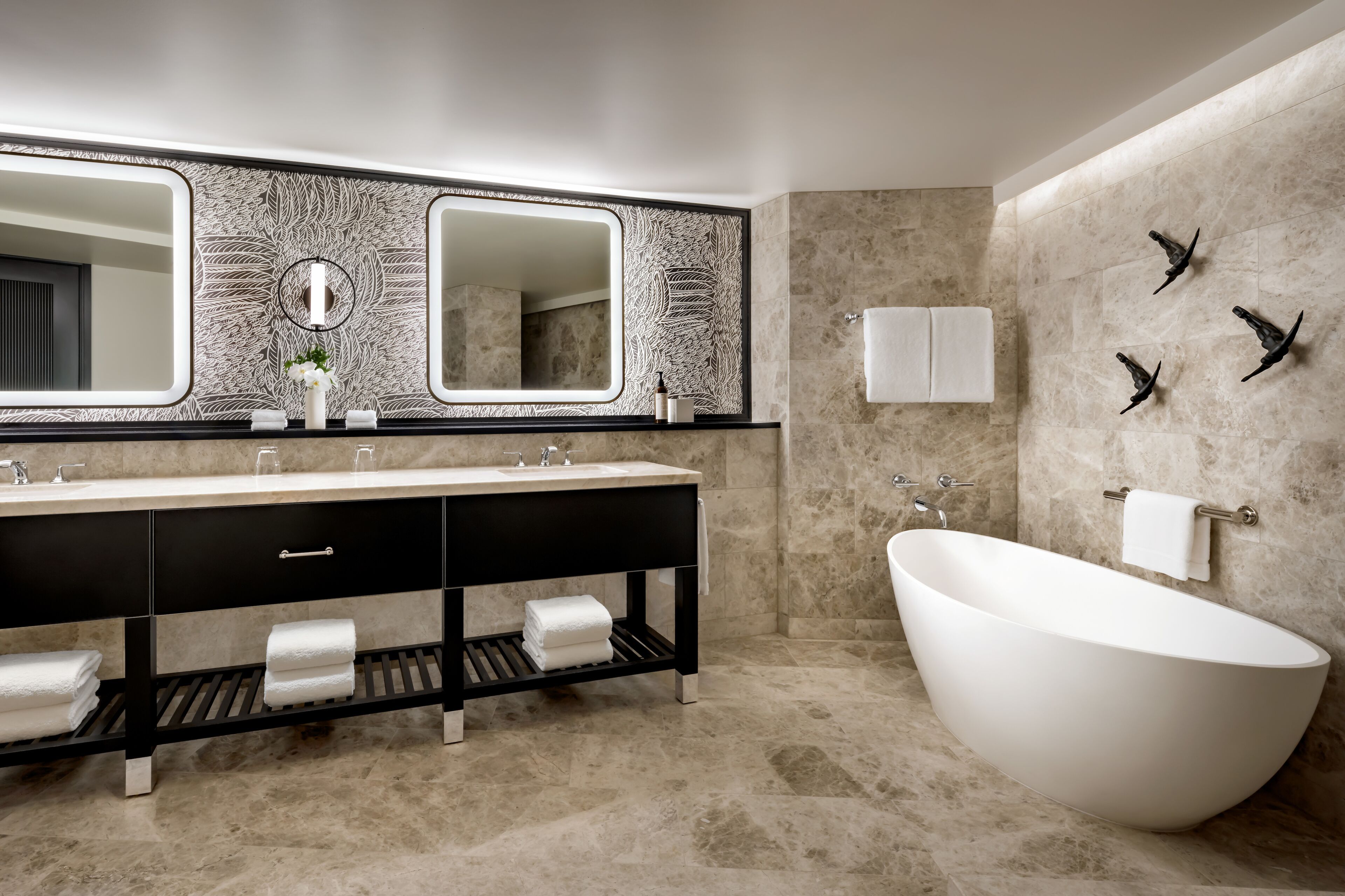 A modern bathroom with a freestanding bathtub, a large mirror with a leaf pattern, and a black vanity with multiple towel holders.