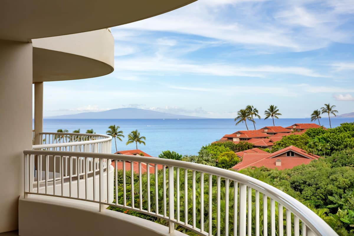A balcony with a view of a coastal area, including houses with red roofs and palm trees.