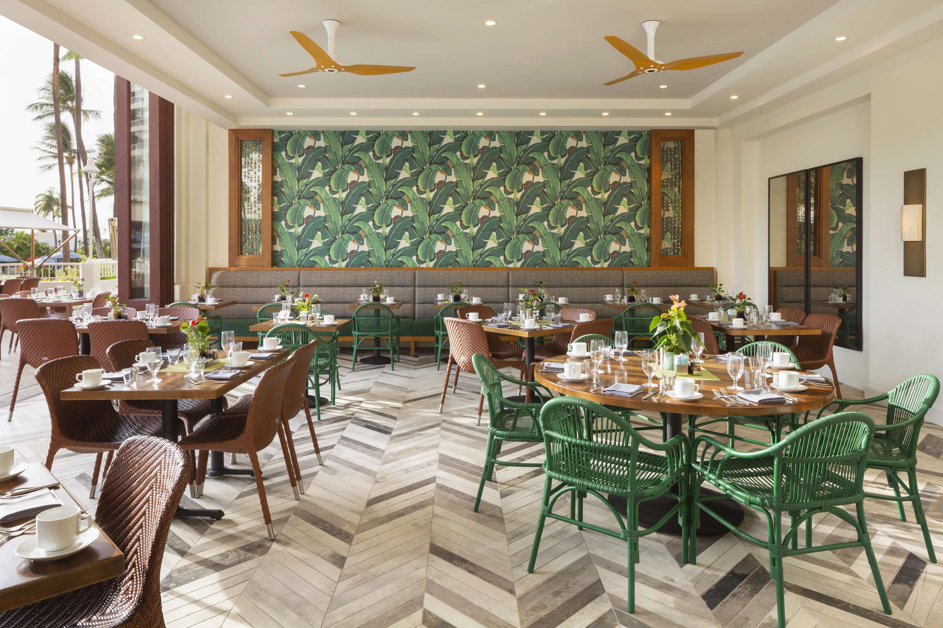 A restaurant with high ceilings, ceiling fans, and a tropical leaf-patterned wall. Tables are set with chairs, and the floor has a herringbone pattern.