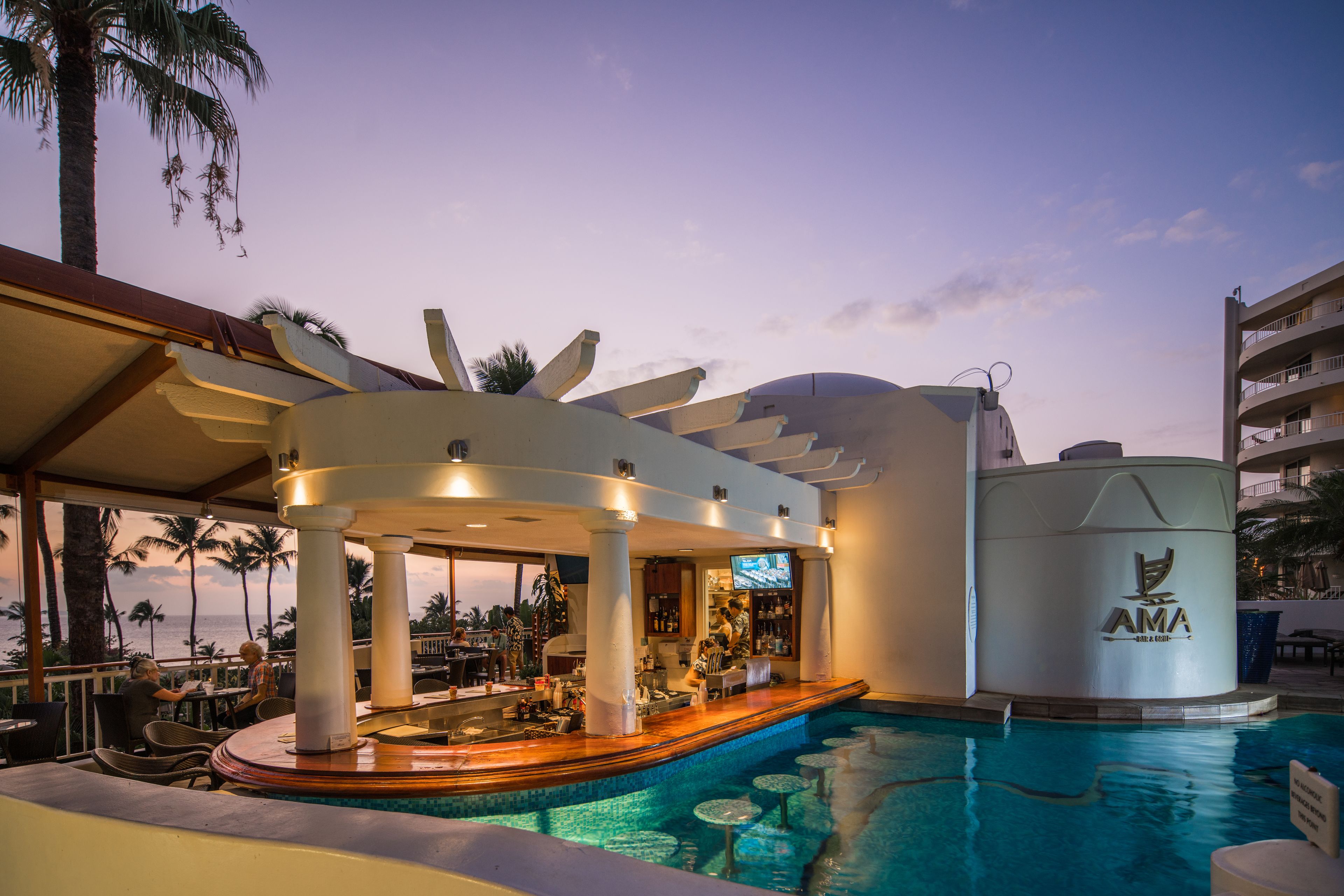 A pool area with a bar, palm trees, and a building with 'AMA' logo.