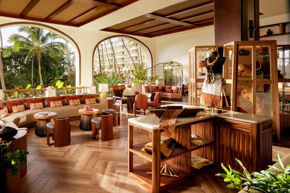 A spacious lounge area with a wooden floor, comfortable seating, and a large window offering a view of palm trees and a building.