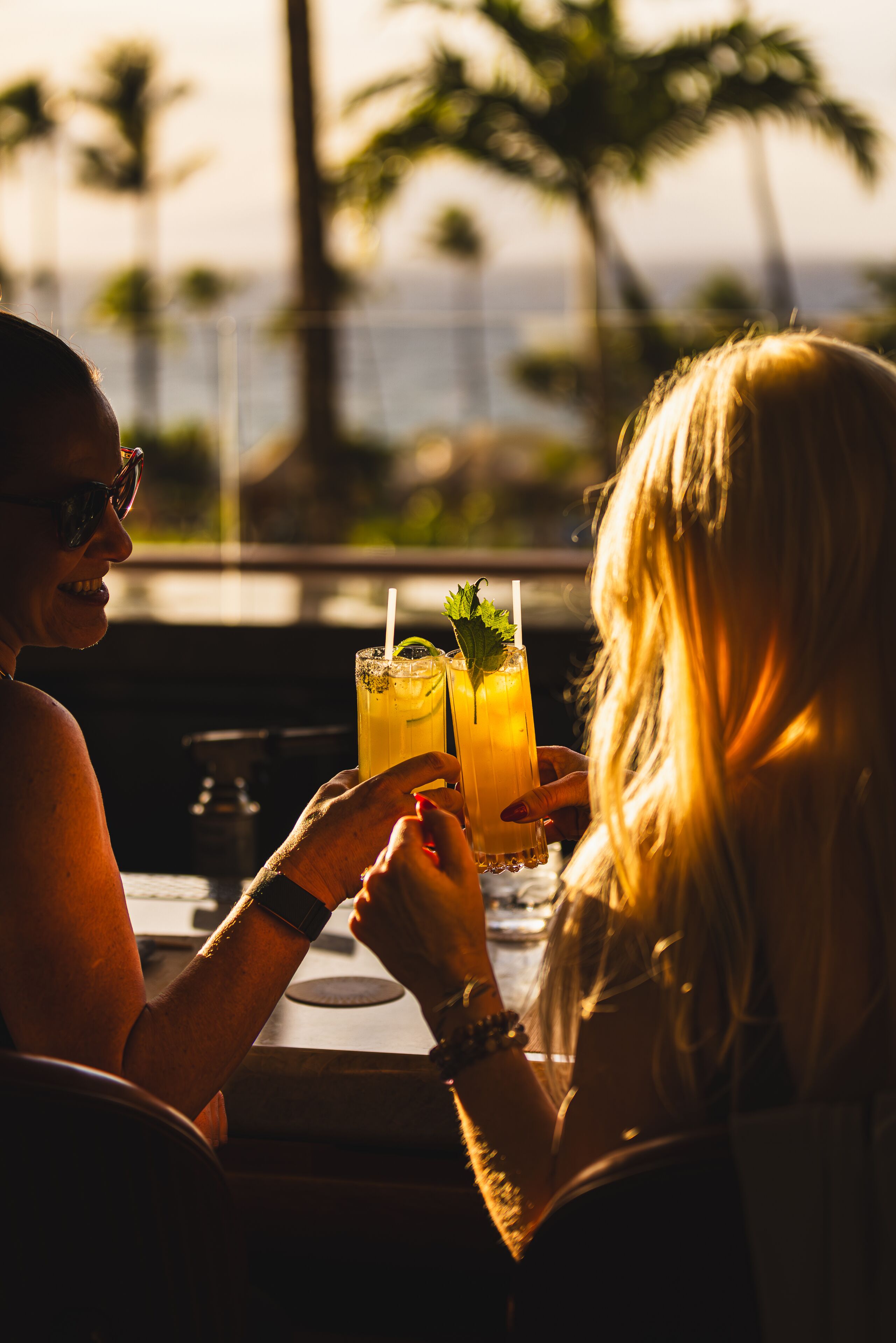 Dos personas sentadas en una mesa con bebidas, con vistas a palmeras y una masa de agua.