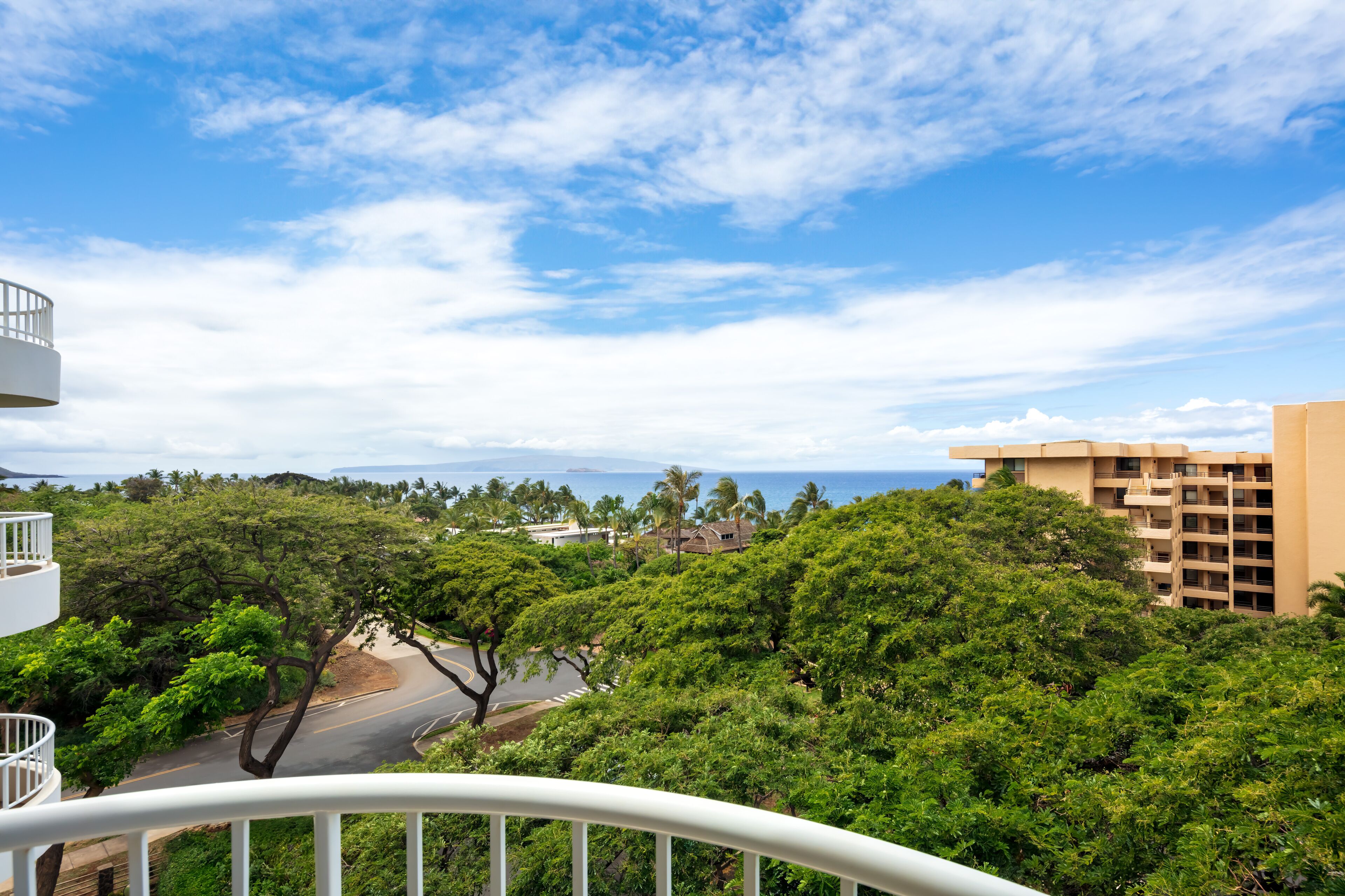 A balcony with a view of a road, lush greenery, and a building in the distance.