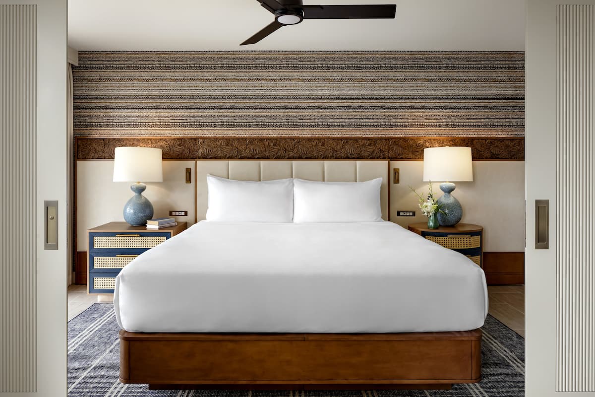 A hotel room with a large bed, two bedside tables with lamps, a ceiling fan, and a textured wall.