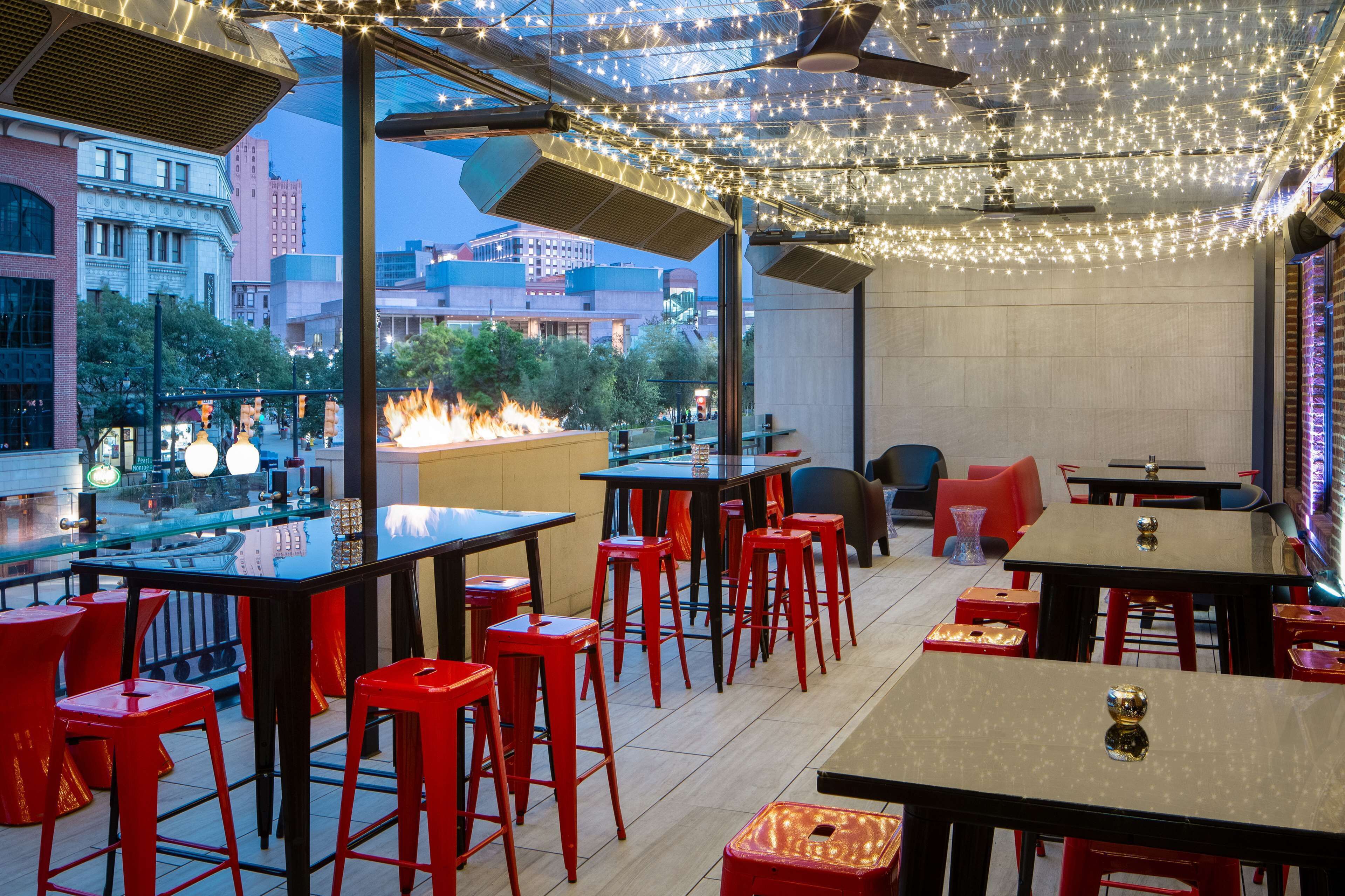 An outdoor dining area with red bar stools and tables, string lights overhead, and a fire feature in the background.