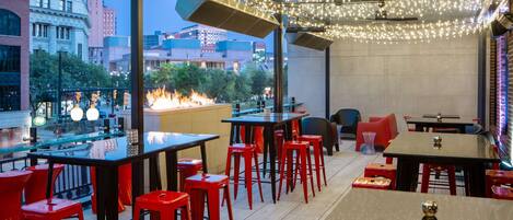 An outdoor dining area with red bar stools and tables, string lights overhead, and a fire feature in the background.