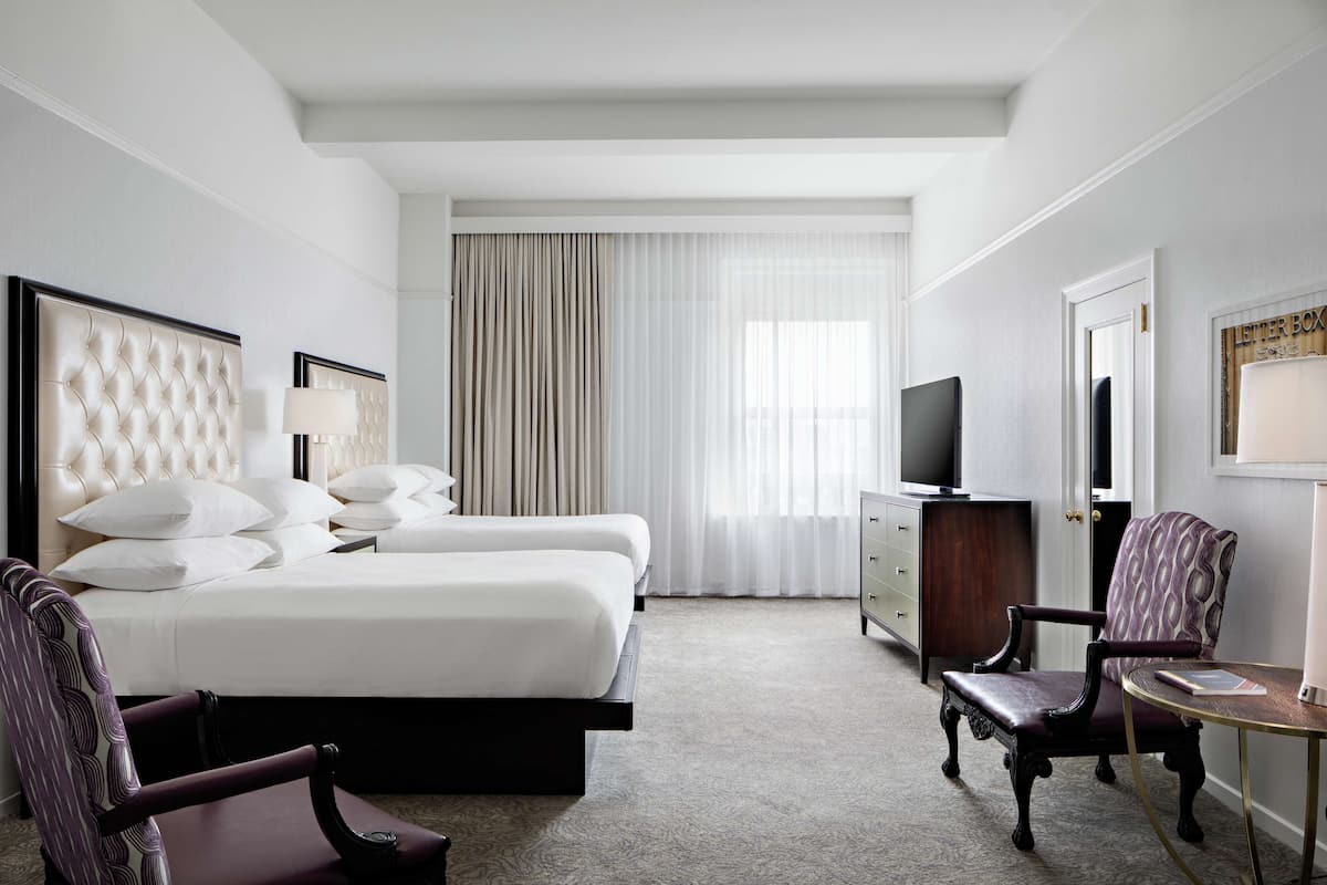 Ein Hotelzimmer mit einem großen Bett, zwei Sesseln, einem Schreibtisch, einem Fernseher und einem Vorhangfenster.