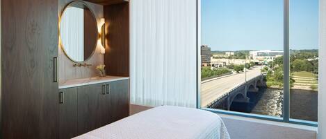 A modern hotel room with a large bed, wooden wardrobe, a round mirror, and a view of a bridge and cityscape.