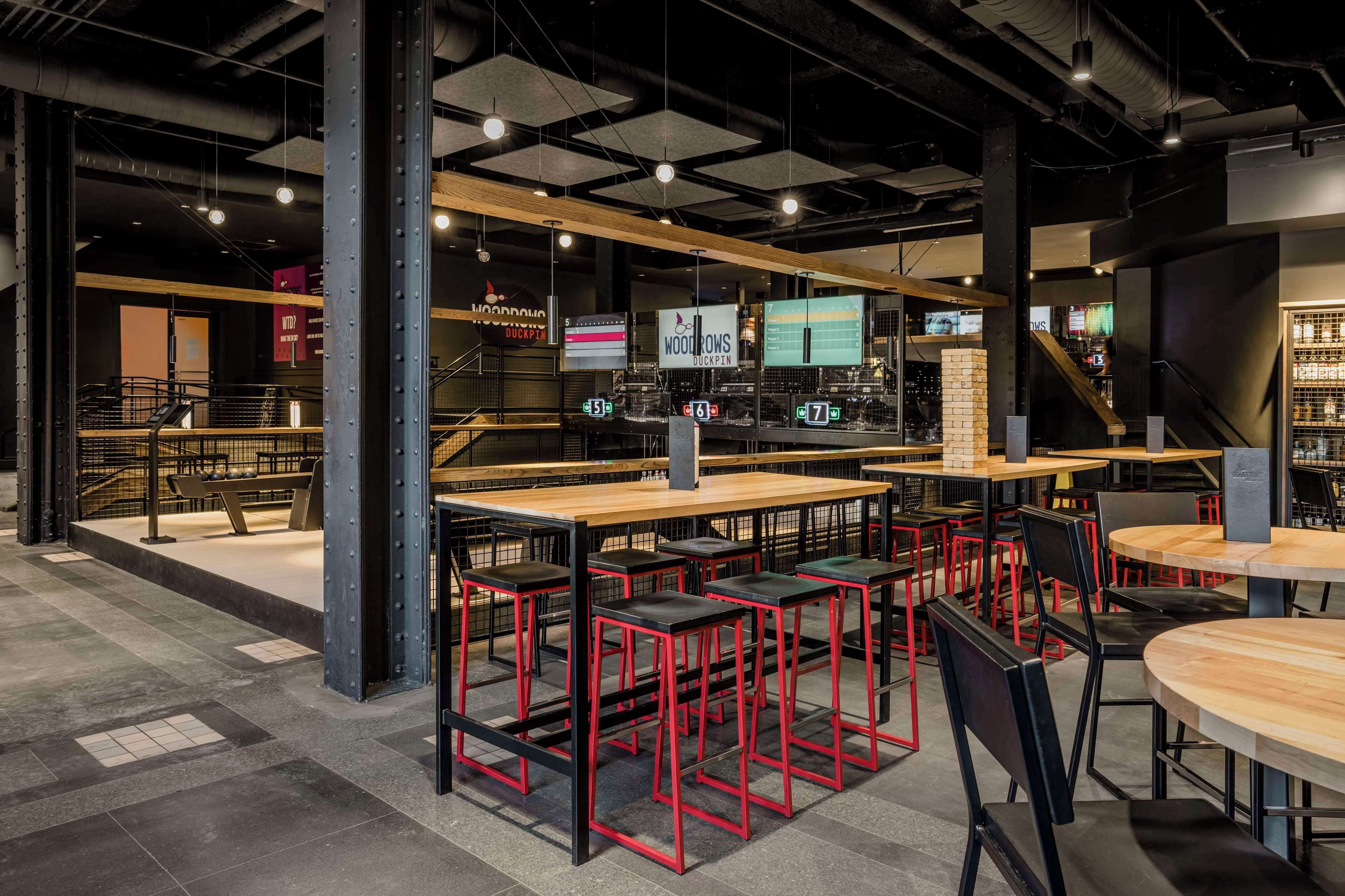 Un espace bar moderne offrant de nombreuses options d’assise, notamment des tables hautes et des tabourets, ainsi qu’un escalier apparent menant à un niveau supérieur.