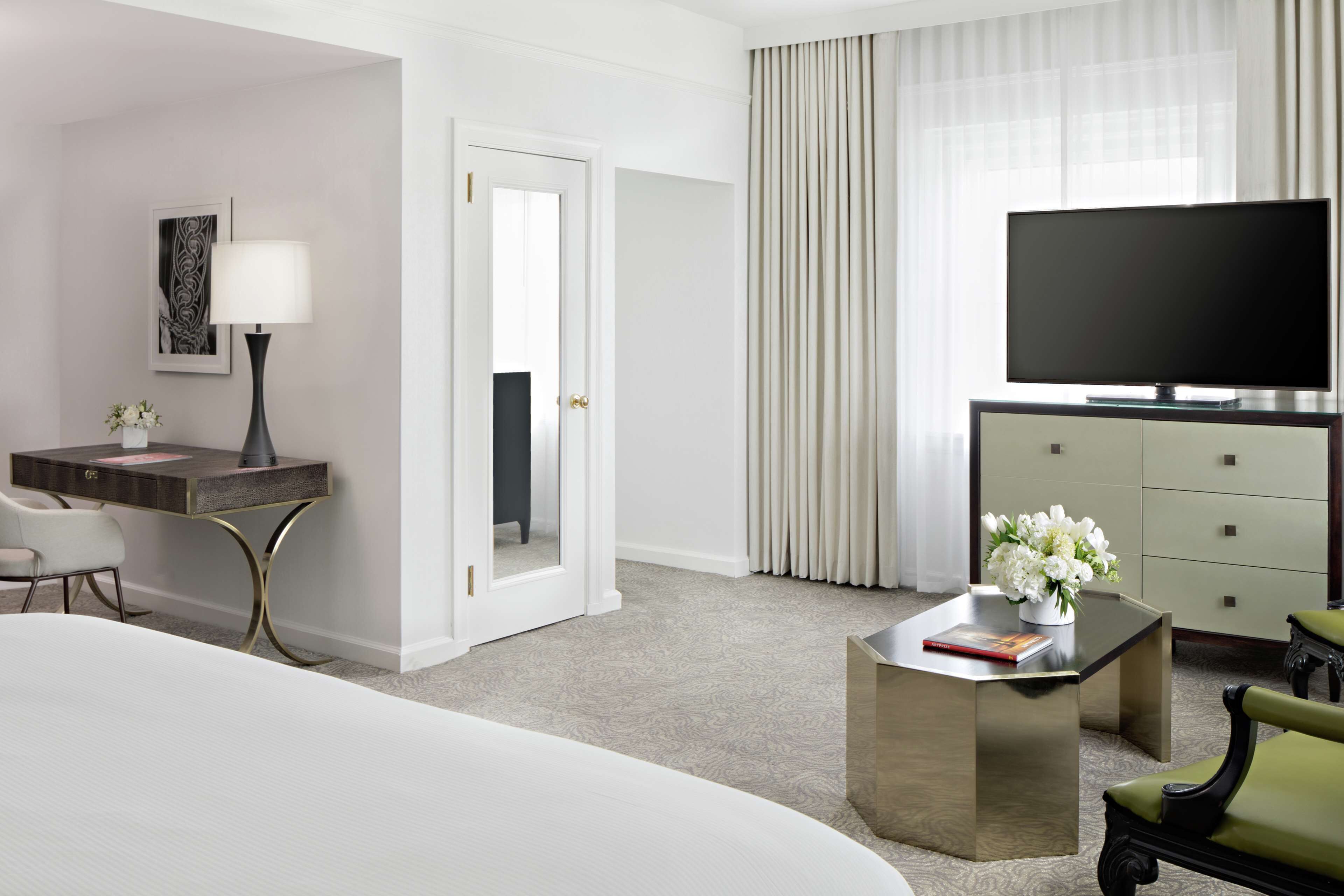 Quarto de hotel com cama, escrivaninha com luminária, televisão e cadeira.