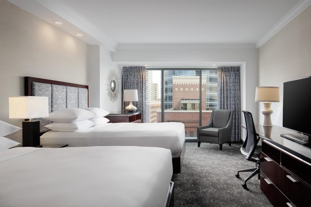 Ein Hotelzimmer mit zwei Betten, einem Schreibtisch, einem Stuhl, einem Fernseher und einem großen Fenster mit Blick auf die Stadt.