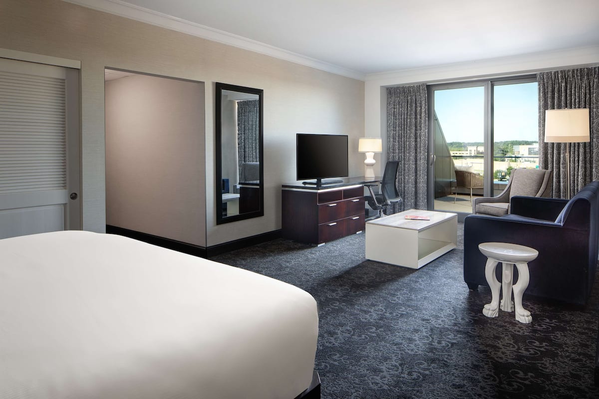 Ein Hotelzimmer mit einem großen Bett, einem Schreibtisch mit Fernseher, einem kleinen Tisch, einer Couch und Blick auf die Stadt durch große Fenster.
