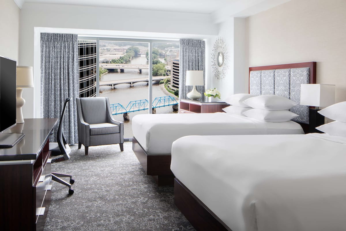 Ein Hotelzimmer mit zwei Betten, einem Schreibtisch, einem Sessel und Blick auf eine Brücke und einen Fluss.
