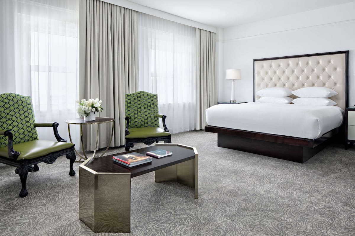 Ein Hotelzimmer mit einem großen Bett, zwei grünen Polsterstühlen, einem kleinen runden Tisch mit einem Buch und einer Blumenvase.