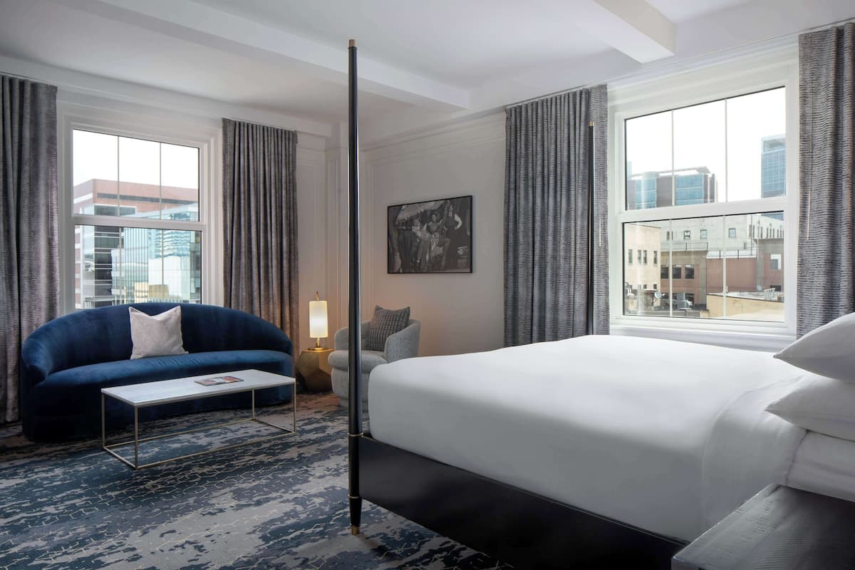Ein Hotelzimmer mit einem großen Bett, einer Couch, einem Couchtisch und Blick auf die Stadt durch große Fenster.