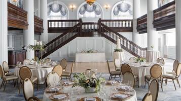 A grand dining hall with multiple round tables set for a formal event, featuring elegant chairs and a central chandelier.