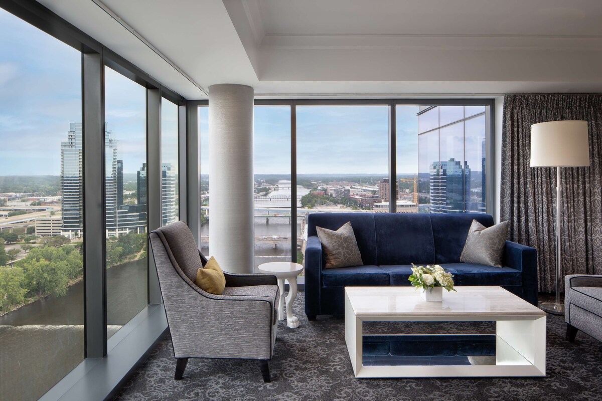 A modern hotel room with a large window offering a city view, a blue sofa, a white coffee table, and a grey armchair.