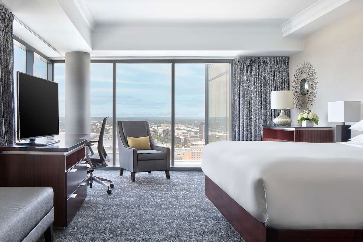 Ein Hotelzimmer mit einem großen Bett, einem Schreibtisch mit Fernseher, einem Sessel und Blick auf die Stadt durch große Fenster.