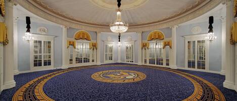 A grand hall with a circular patterned carpet, ornate chandeliers, and classical architecture.