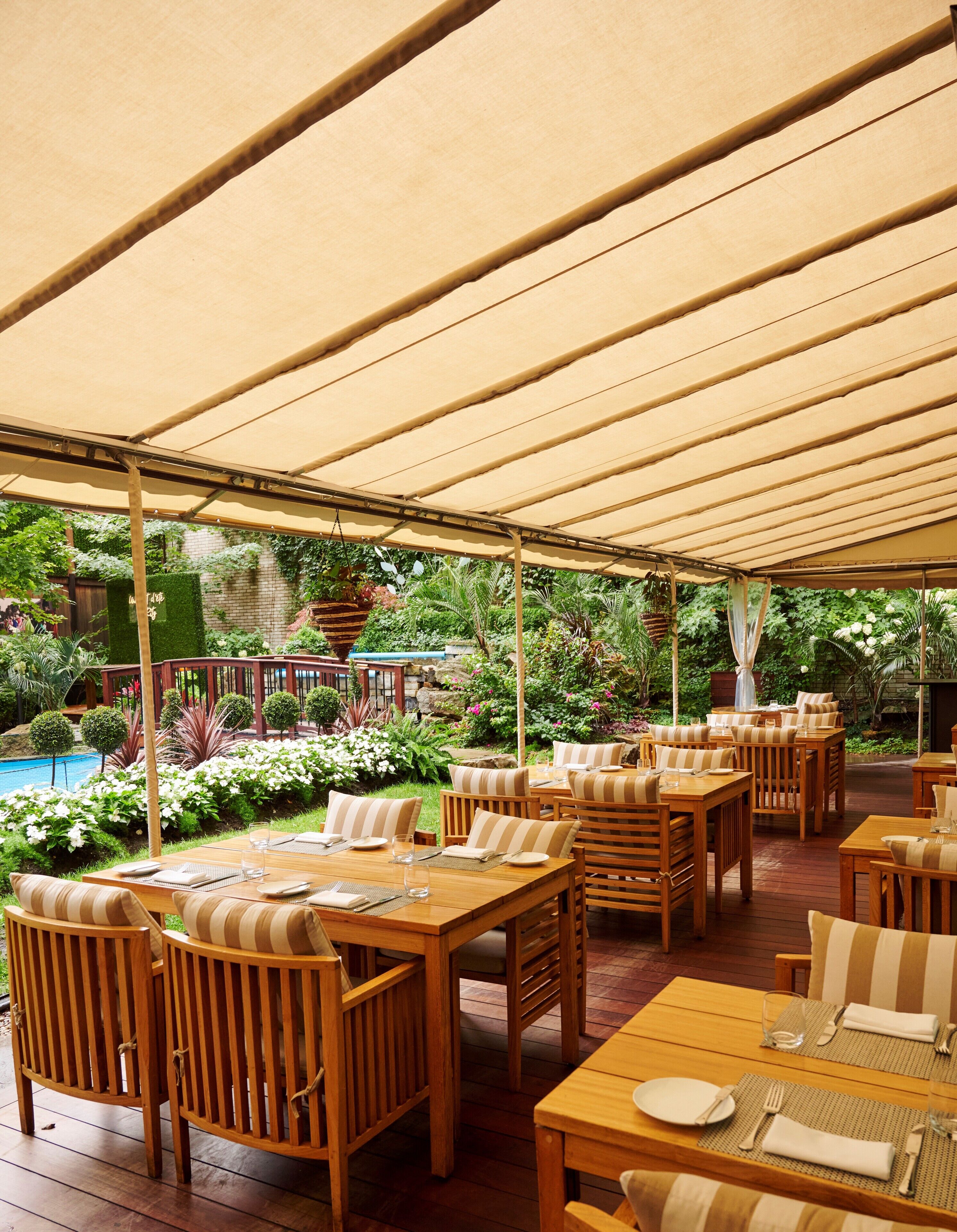 Un patio al aire libre con mesas y sillas de madera, una piscina y abundante vegetación.