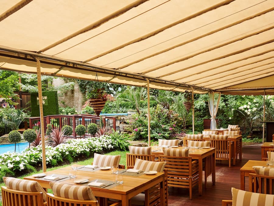 Un patio al aire libre con mesas y sillas de madera, una piscina y abundante vegetación.