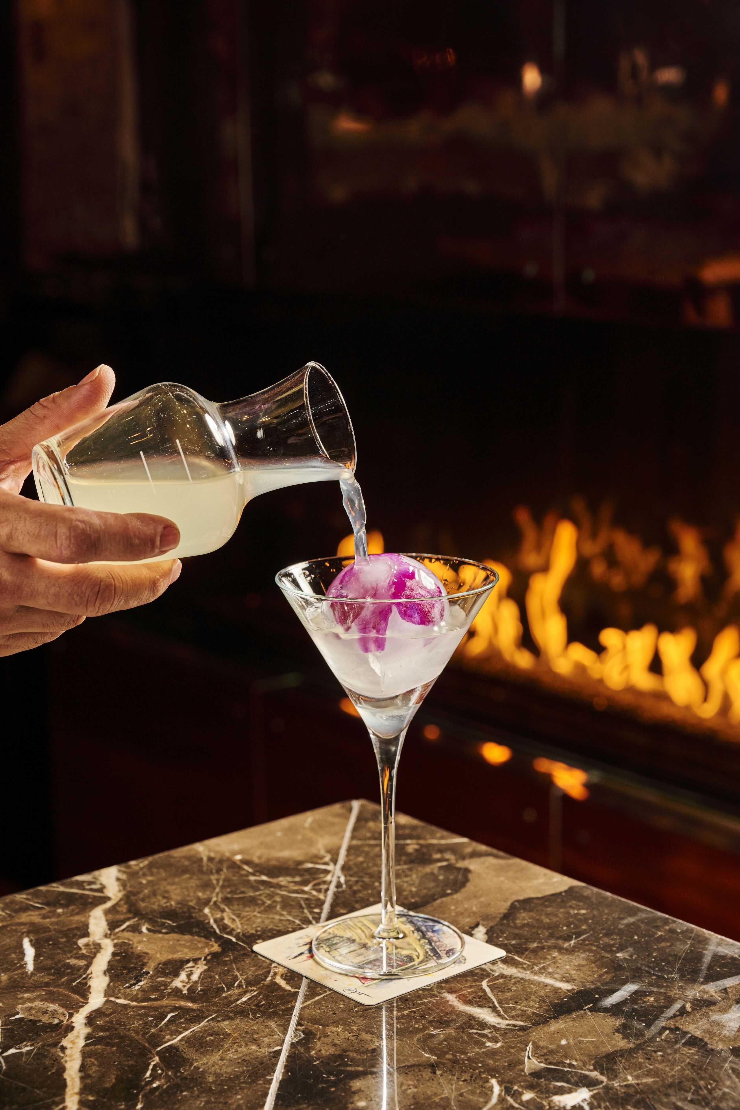 Een hand giet een heldere vloeistof in een martini-glas met ijsblokjes op een marmeren aanrecht.