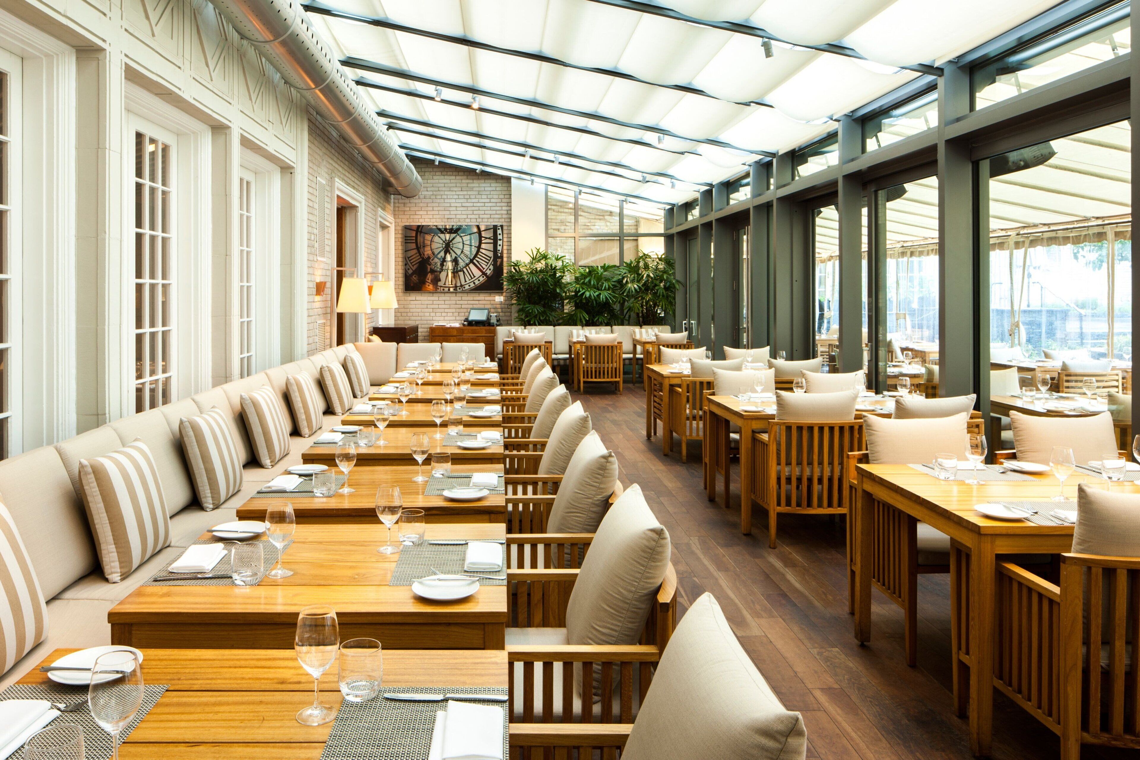 A spacious restaurant with long wooden tables and cushioned seating, large windows, and a brick accent wall.