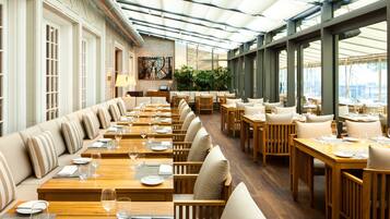 A spacious restaurant with long wooden tables and cushioned seating, large windows, and a brick accent wall.