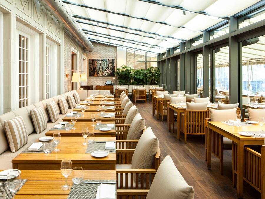 A spacious restaurant with long wooden tables and cushioned seating, large windows, and a brick accent wall.