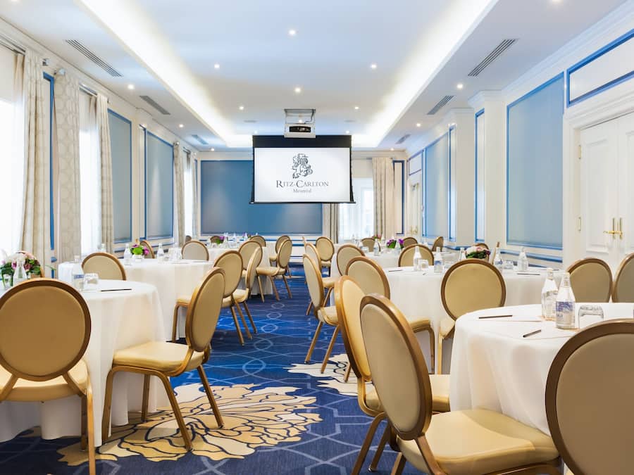 Un salón de conferencias con filas de mesas y sillas, una pantalla grande al frente y una alfombra azul.