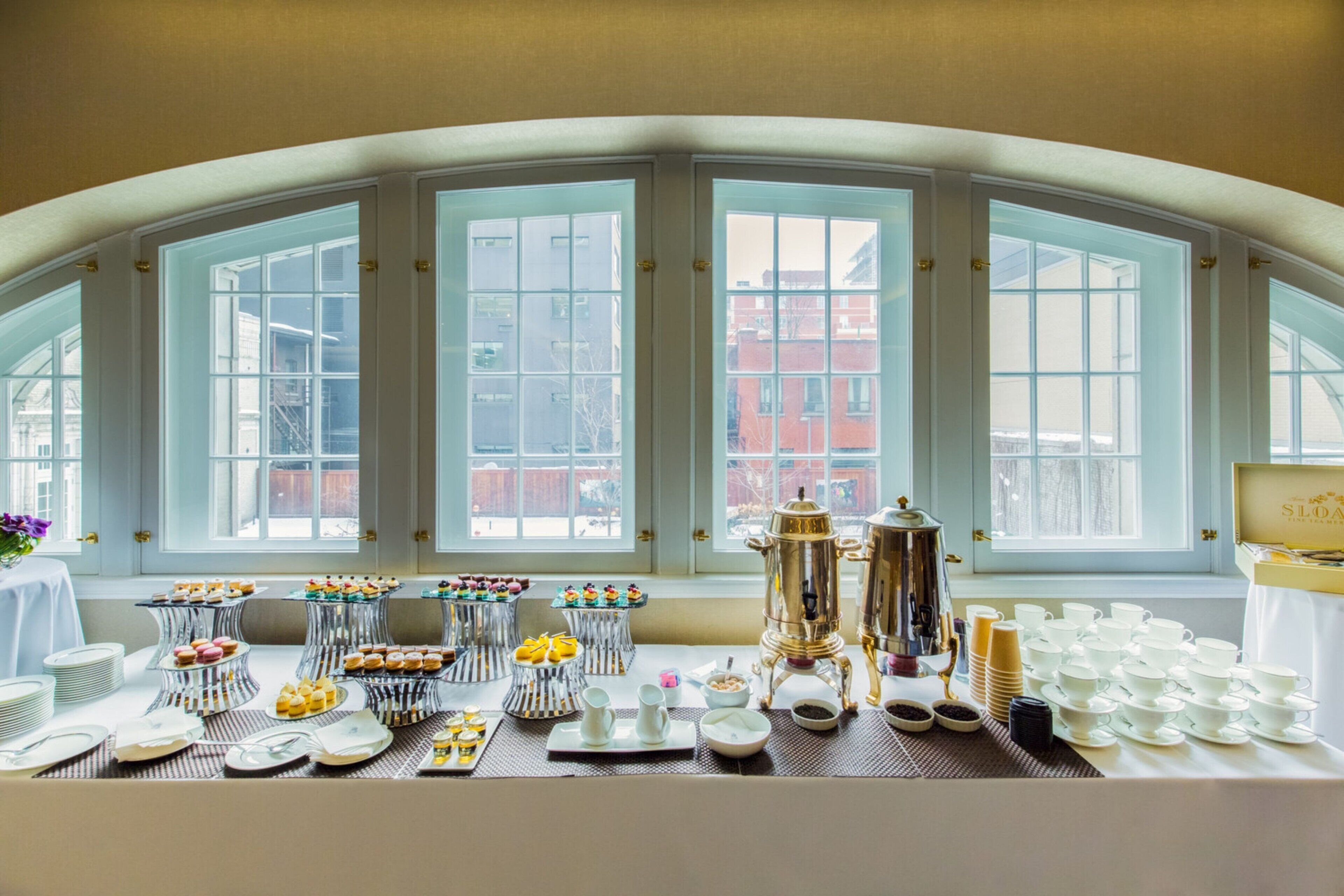 A breakfast buffet with various pastries and beverages, set against a backdrop of large windows with a view of buildings.