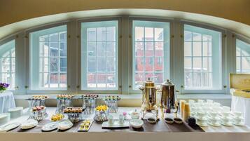 A breakfast buffet with various pastries and beverages, set against a backdrop of large windows with a view of buildings.