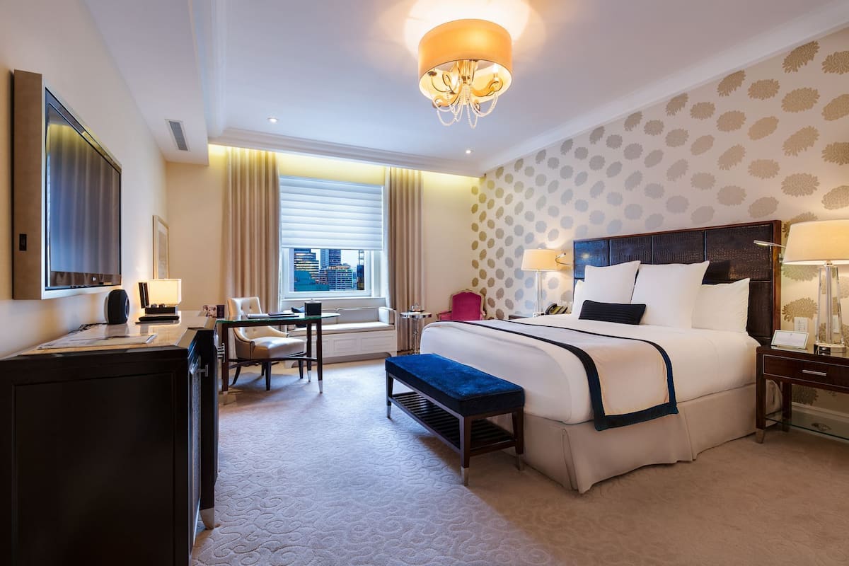 A hotel room with a large bed, a desk with a chair, a small table, a bench, and a window with a city view.